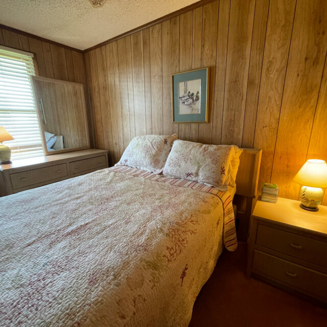 Cozy bedroom with wood-paneled walls, a double bed, two lamps, a dresser with mirror, and a picture above the bed.