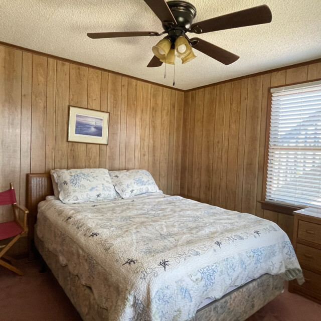 Small bedroom with wood-paneled walls, a bed, director’s chair, dresser, ceiling fan, and window with blinds.