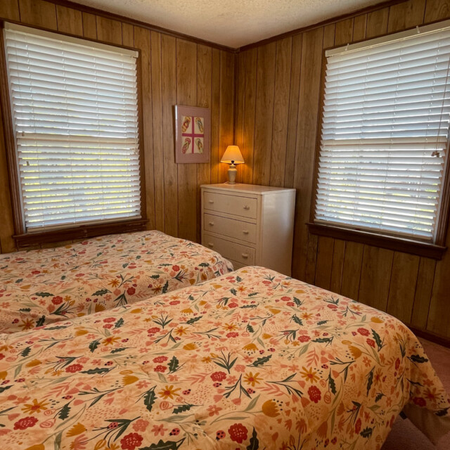 Bedroom with two twin beds, floral bedding, wood-paneled walls, two windows, dresser, and table lamp.