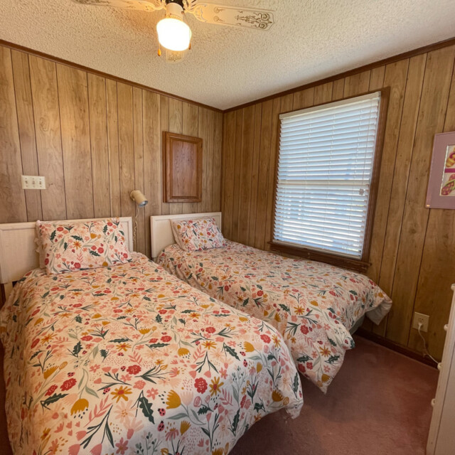 Small bedroom with two twin beds, floral bedding, wood-paneled walls, window with blinds, and dresser.