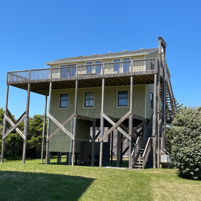 Elevated beach house on stilts with a large deck, outdoor stairs, and grassy yard under a clear blue sky.