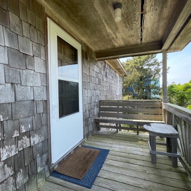 Small wooden porch with a bench, round table, doormat, shingled wall, and a white door under a weathered roof.