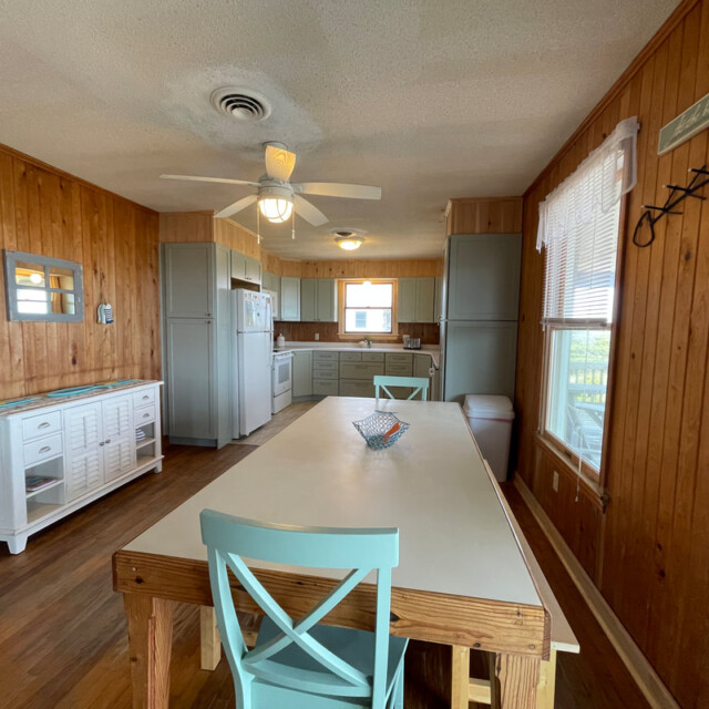 Wood-paneled kitchen with white cabinets, a dining table, blue chairs, ceiling fan, and sunlight through windows.