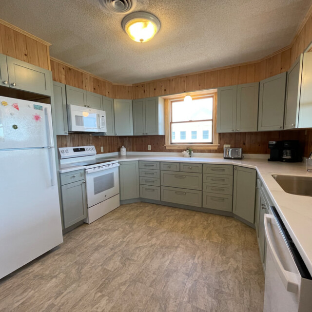 L-shaped kitchen with light green cabinets, white appliances, wood paneling, and a window above the sink.