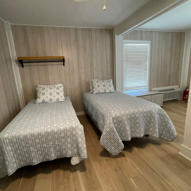 Two twin beds with patterned bedding in a simple, wood-paneled room with light wood flooring and windows.