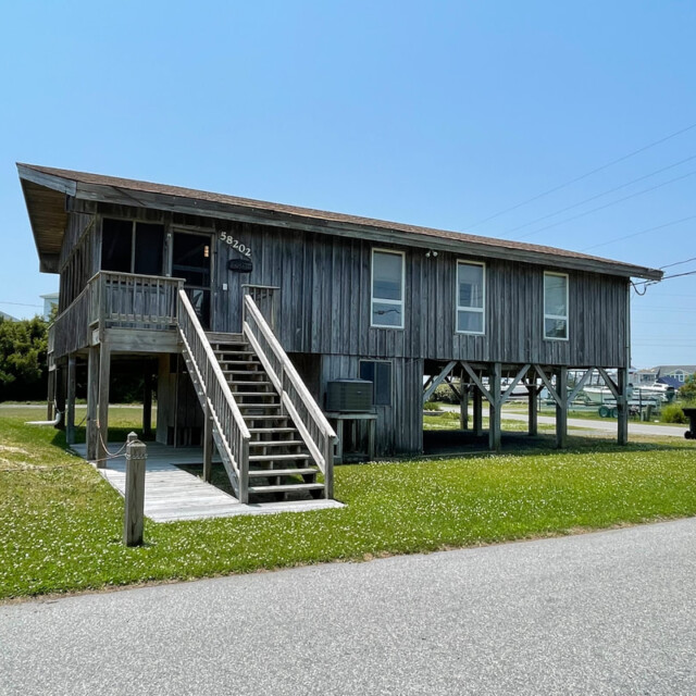 Wooden elevated beach house with stairs, grass lawn, and paved road under a clear blue sky.