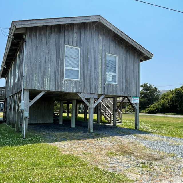 A weathered wooden house on stilts with stairs, gravel driveway, and grassy yard under a clear blue sky.