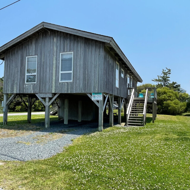 A small gray wooden house on stilts with stairs, a gravel driveway, and a rental sign in the front yard.