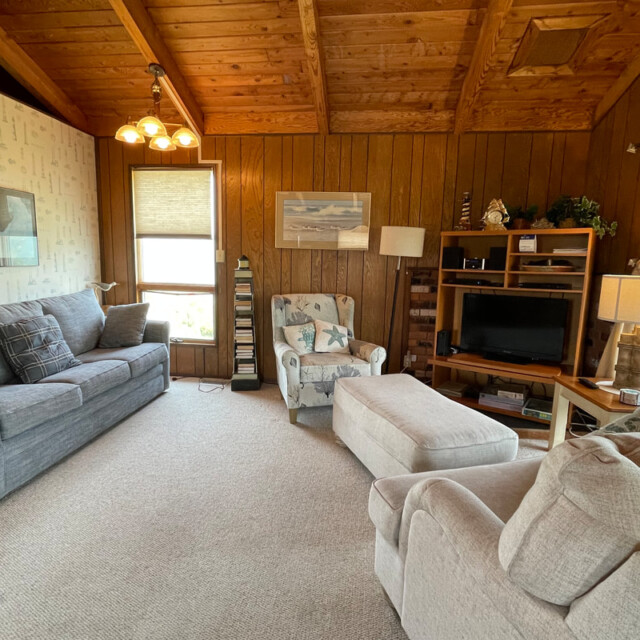 Cozy living room with wood paneling, gray sofas, an armchair, a TV stand, and large windows with beige curtains.