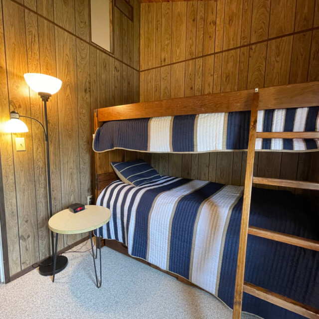 Wood-paneled room with a wooden bunk bed, striped bedding, a floor lamp, and a small round table with an alarm clock.