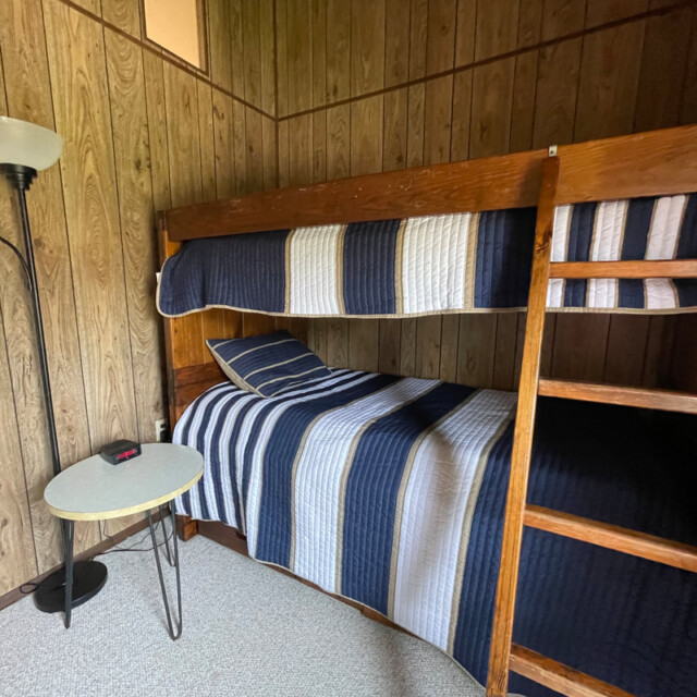 Wood-paneled room with a bunk bed, blue and white striped bedding, a small round table, lamp, and clock.