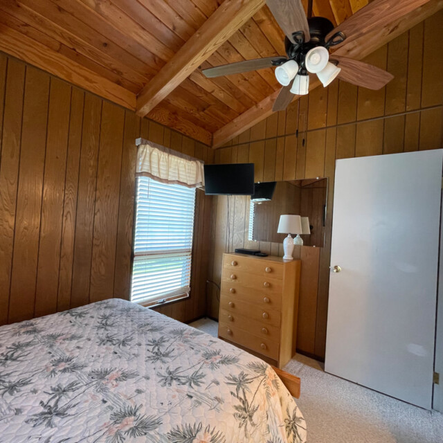 Wood-paneled bedroom with a bed, dresser, TV, ceiling fan, window with blinds, and an open door.