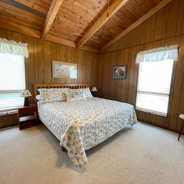 Bedroom with wood-paneled walls, a king bed with floral bedding, two windows, and a wall-mounted TV.
