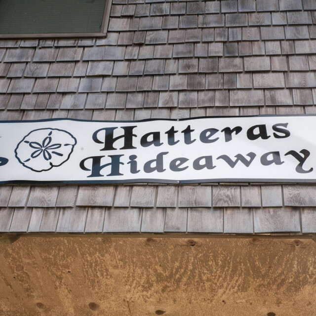 A wooden sign reads “Hatteras Hideaway” with a sand dollar illustration, mounted on a shingled wall.