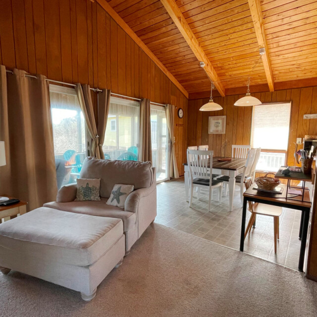 Living room with beige chaise lounge, wooden walls, dining table, and large windows letting in natural light.