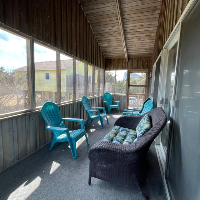 Screened porch with wicker loveseat, blue plastic chairs, and view of a yellow house outside.