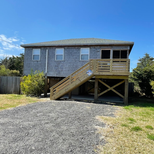A gray wooden house on stilts with a front porch, ramp, stairway, gravel driveway, and surrounding shrubs.