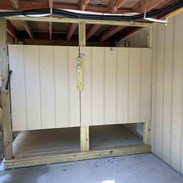 Outdoor wooden enclosure with a lock under a raised roof, adjacent to a fence and siding, on a concrete pad.