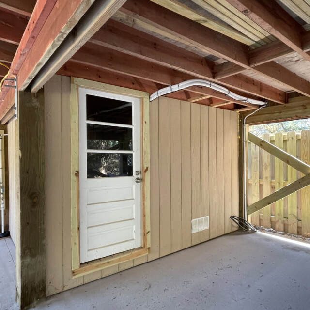 A wooden door with three glass panes set in a tan wall under a covered patio next to a wooden fence.