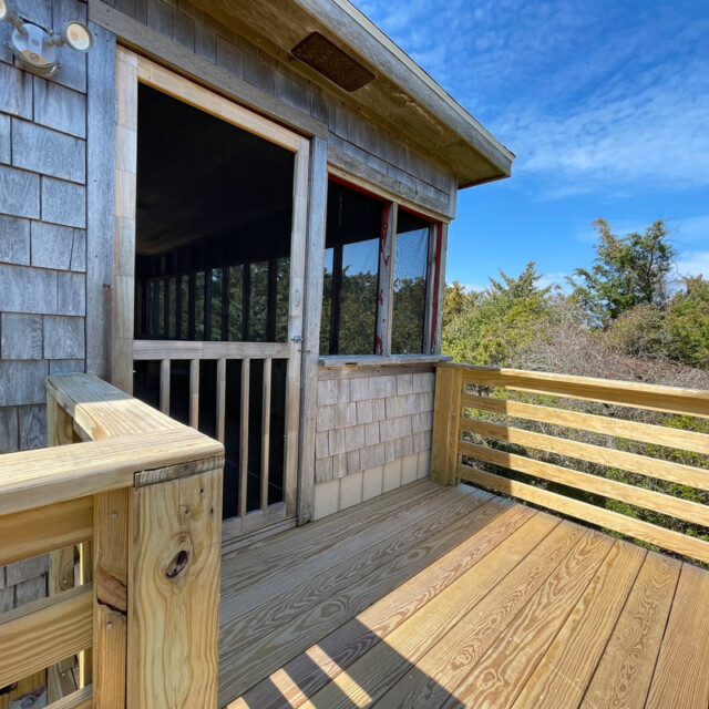 Wooden deck with railing attached to a shingled house, next to a screened porch and trees under a blue sky.