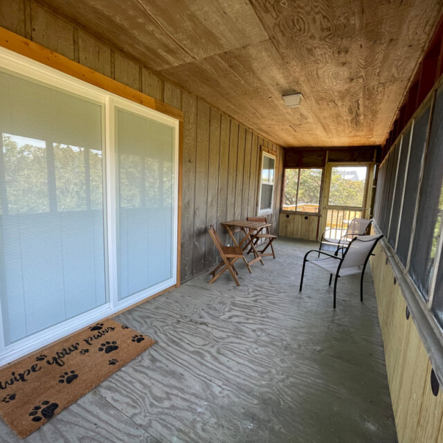 Screened-in porch with sliding glass door, small table and chairs, metal chair, and a welcome mat on concrete floor.