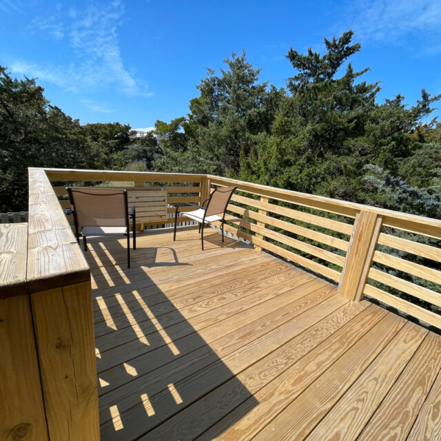 Wooden deck with two chairs and a table, surrounded by trees under a clear blue sky.