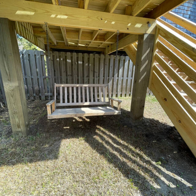 Wooden porch swing hanging under a deck next to a wooden staircase and a fenced backyard area on a sunny day.