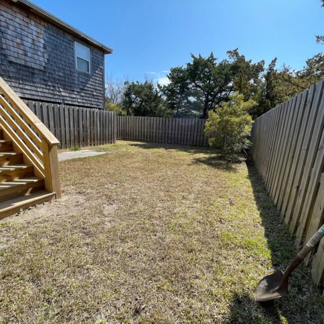 Fenced backyard with a wooden staircase, a small tree, and a shovel stuck in the grassy ground on a sunny day.