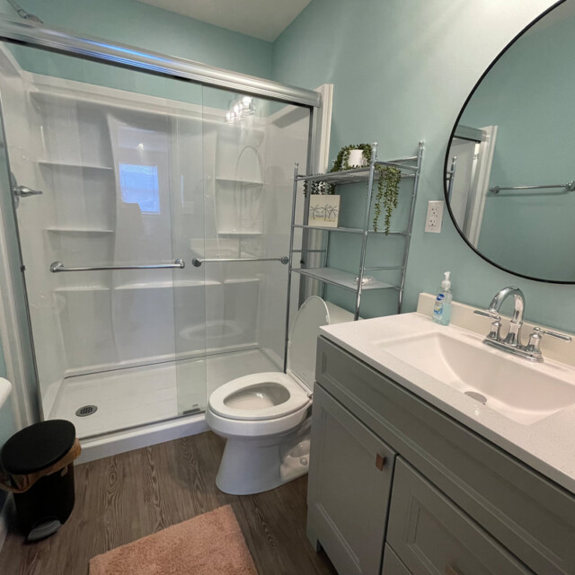 Bathroom with a glass shower, toilet, vanity with sink, round mirror, metal shelf, and light blue walls.