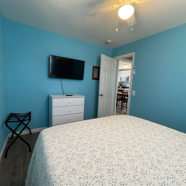 Small bedroom with blue walls, a bed, a dresser, wall-mounted TV, and an open door leading to a kitchen area.