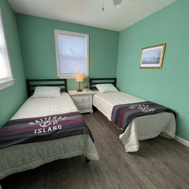 Two twin beds with “Hatteras Island” blankets in a green-walled room with a nightstand, lamp, and wall art.