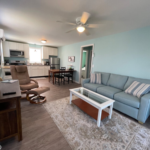 Living room with blue sofa, glass coffee table, recliner, TV, and open kitchen in background with dining table.