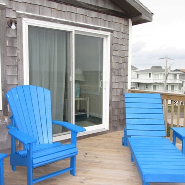 Two blue wooden chairs and a table are on a wooden deck outside a house with gray siding and a sliding glass door.