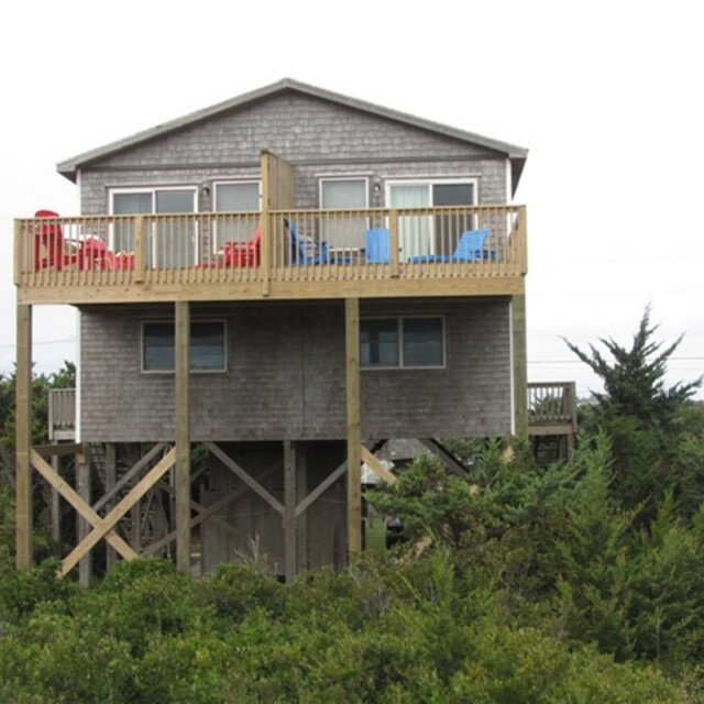 Two-story wooden beach house on stilts with a balcony, red and blue chairs, surrounded by green shrubs and trees.