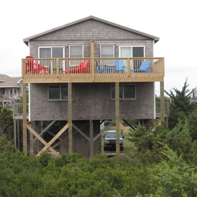 Two-story beach house on stilts with a wooden deck, colorful chairs, and a car parked underneath, surrounded by greenery.