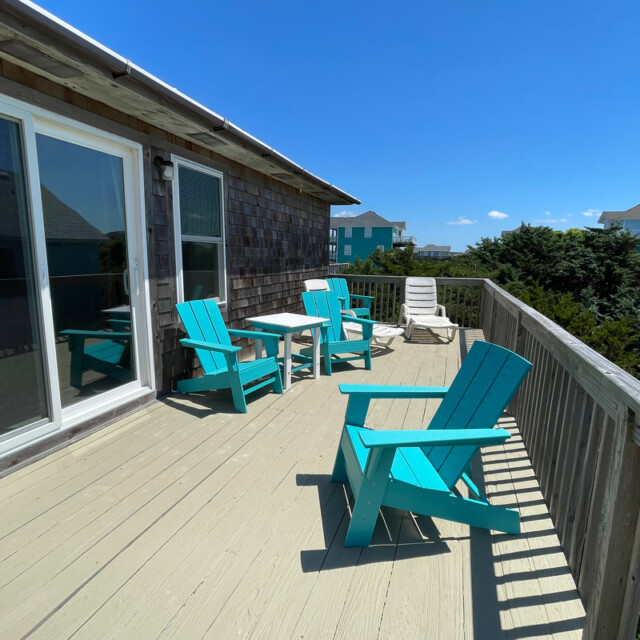 Wooden deck with turquoise chairs and a table, overlooking greenery and nearby houses under a clear blue sky.