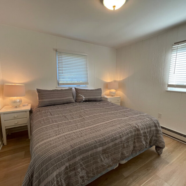 Bedroom with a king-size bed, two nightstands with lamps, wood floor, and two windows with blinds.