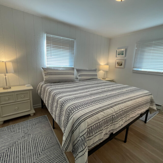Bedroom with a striped bed, two nightstands with lamps, rug, wood floors, and two windows with blinds.