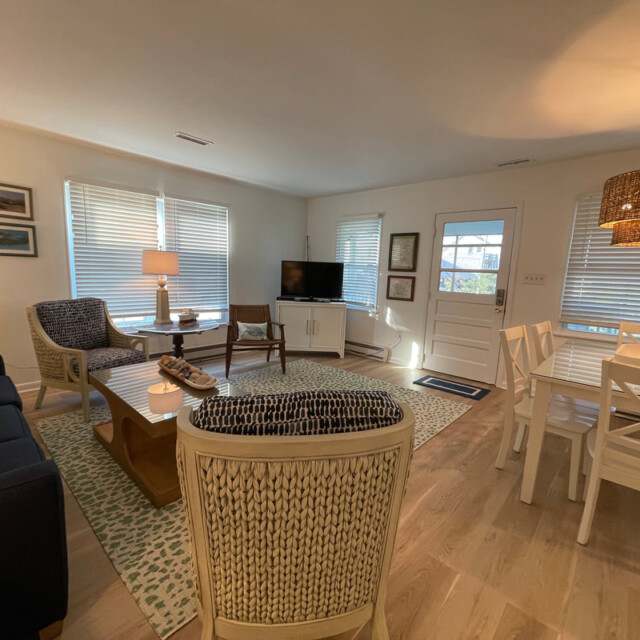 Bright living and dining area with woven chairs, white table, TV, art on walls, and large windows with blinds.