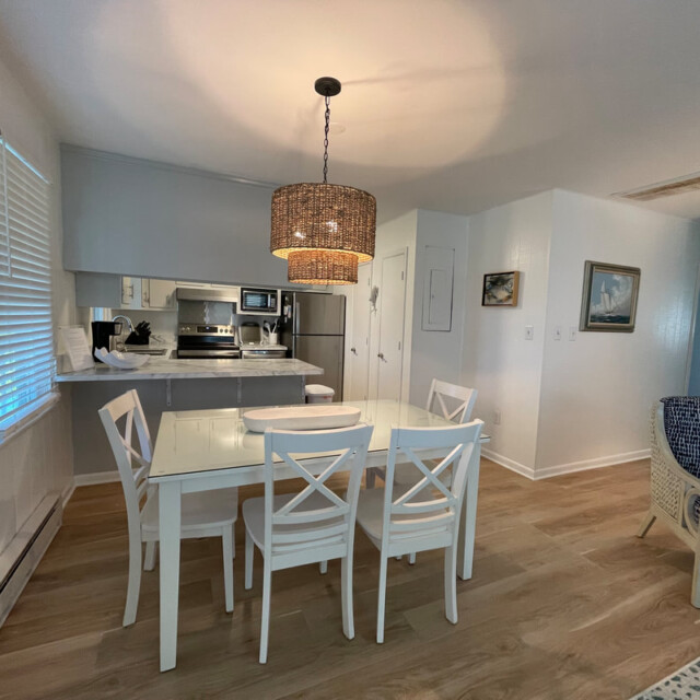 A modern kitchen and dining area with a glass table, white chairs, pendant light, and light wood flooring.