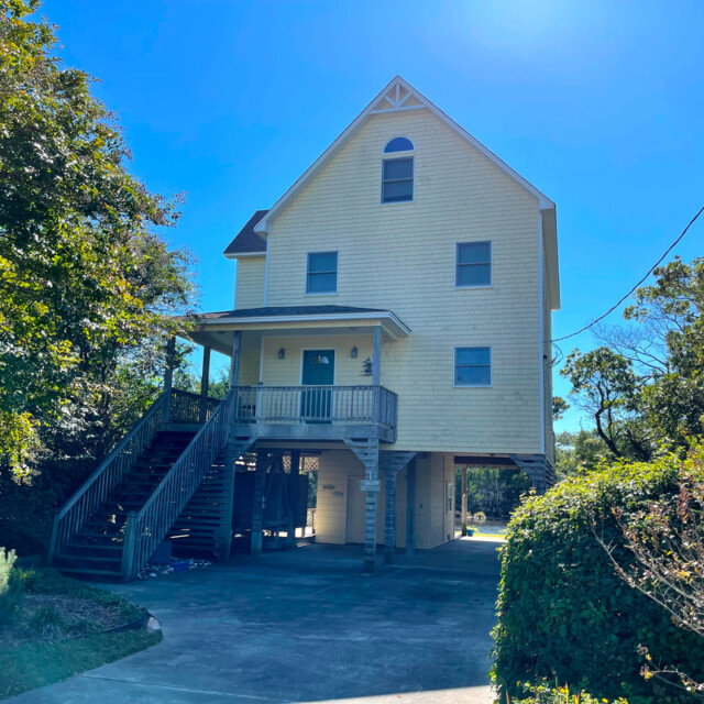 Three-story yellow house on stilts with a front porch, exterior stairs, and a driveway, surrounded by trees and bushes.