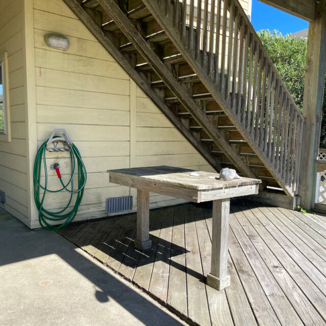 Wooden table and hose on a deck under outdoor stairs beside a beige building on a sunny day.