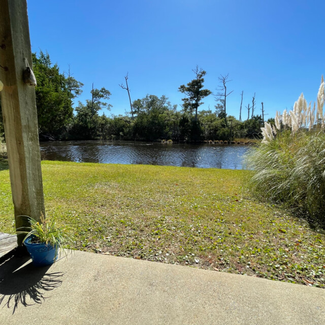 View of a grassy yard with a potted plant, overlooking a calm pond and trees under a clear blue sky.