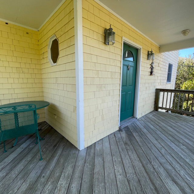 Wooden porch with yellow shingle siding, a green table and chairs, green door, and two wall lanterns.
