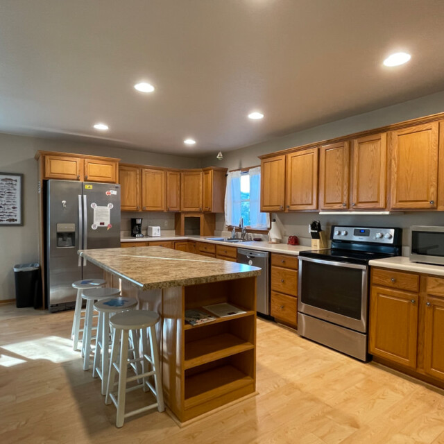 Spacious kitchen with wooden cabinets, stainless steel appliances, island with stools, and bright overhead lighting.