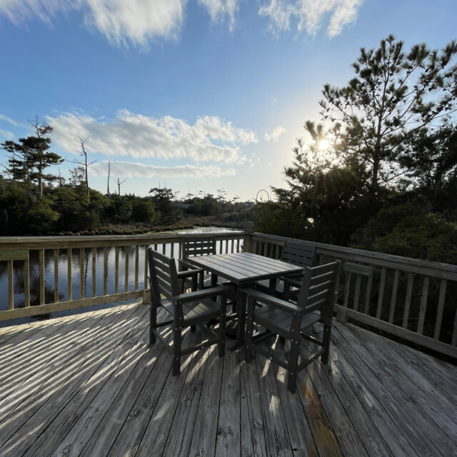 Wooden deck with a table and six chairs overlooks a calm river, surrounded by trees under a partly cloudy sky.