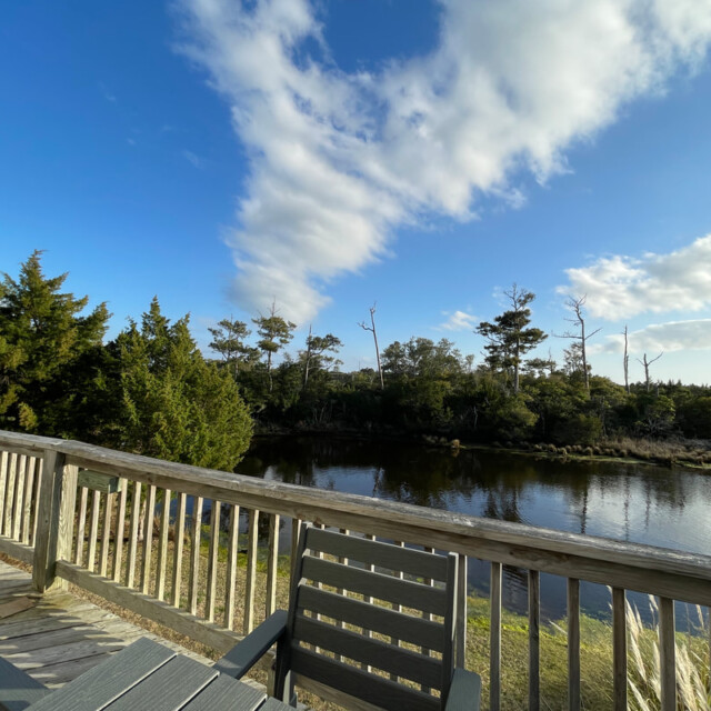 Wooden deck with chairs overlooking a pond, surrounded by trees under a blue sky with scattered clouds.