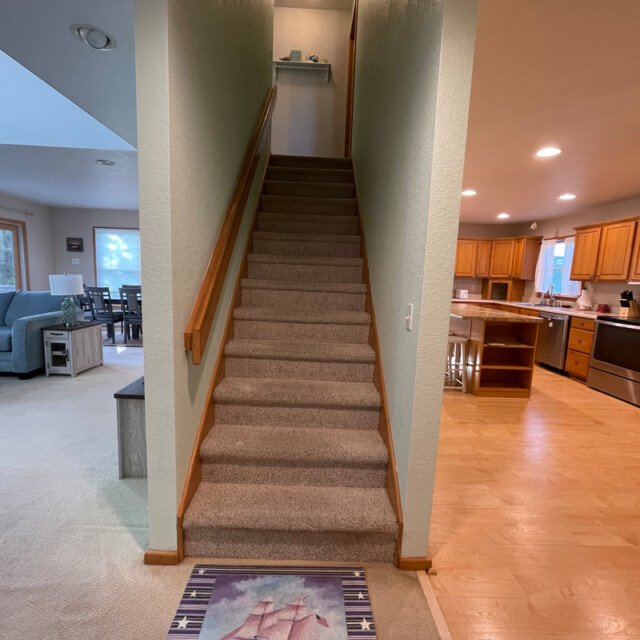 Carpeted stairs between a living room with blue sofas and a kitchen with wooden cabinets and light wood flooring.