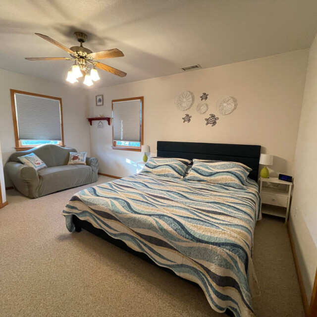 Bedroom with a double bed, patterned bedding, armchair, ceiling fan, and wall decor on a beige carpeted floor.