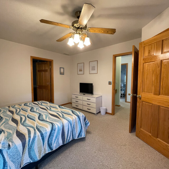 Bedroom with a bed, ceiling fan, dresser with TV, and open doors to hallway and bathroom.